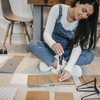 woman with long hard hair wearing white long sleeves and blue overalls drills through tiles on the floor