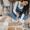 16 woman with long hard hair wearing white long sleeves and blue overalls drills through tiles on the floor