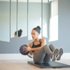 Biracial woman wearing black sports bra and grey workout pants using medicine ball while exercising abs on black workout mat in front of mirrors