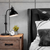 Close up of light rustic wooden bedside table with a black bendable lamp and alarm clock. On right is black headboard queen bed with white stripes sheets and black furry accent pillows.