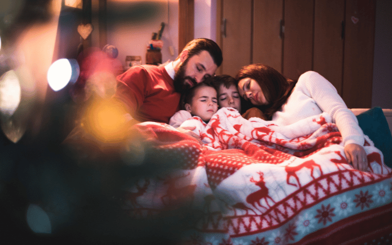 Husband and wife and two kids snuggled up under red and white Christmas blanket on couch