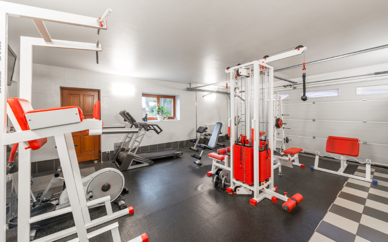 Garage gym with black rubber flooring over checkerboard tiles. Exercise machines are orange and white.