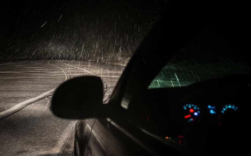 dark night scene with dark sedan vehicle driving down poorly lit road in blowing snow storm