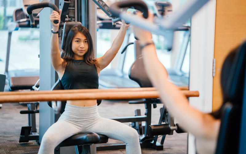young woman with dark hair using an arm exercise machine in front of a mirror, GYM MEMBERSHIP OR HOME GYM? IN 2023 wearing a black tank top and light grey workout pants