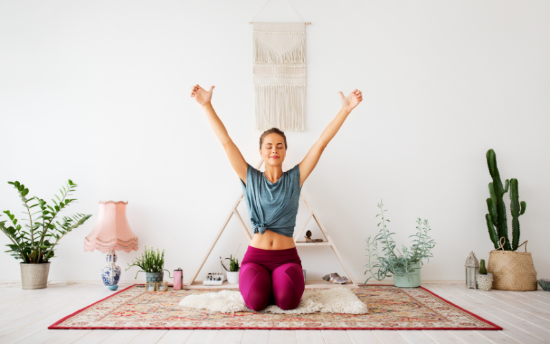 Caucasian woman wearing blue t-shirt and magenta workout pants kneeling making V with her arms on red patterned carpet in front of pyramid shaped white shelf in plant room
