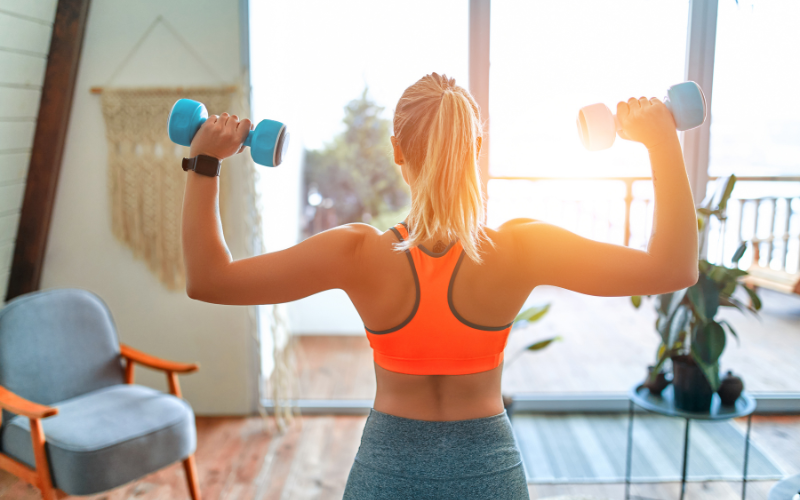 Caucasian woman wearing bright orange sports bra from back lifting light blue hand weights in front of bright open glass double doors in home gym beside velvet blue and wooden chair 
