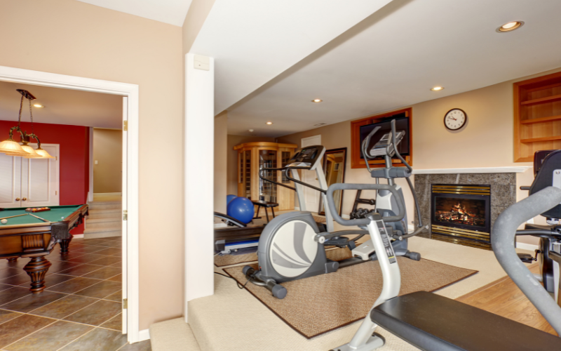 Home gym in front of elevated area by fire place in basement beside billiard room with exercise bike, squat rack and dark blue pilates ball