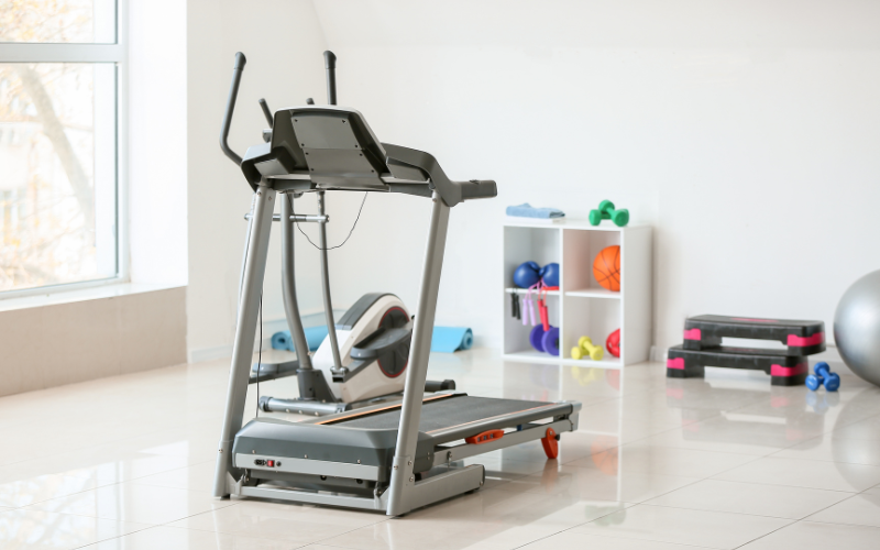 Grey treadmill and exercise bike in bright, lit, white home gym over tiled floor with neon free hand weights on white shelf in back ground beside black and pink step up steps and grey pilates ball