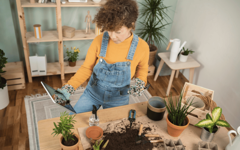 Female YouTuber repotting at her desk while holding ipad wearing long sleeved yellow tshirt and light blue overalls