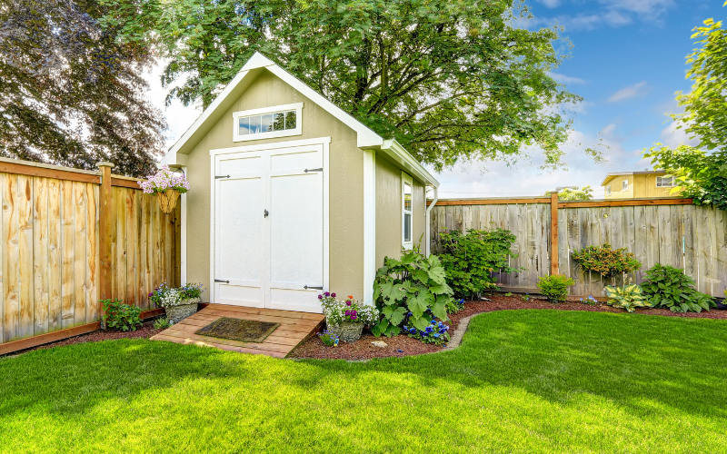 Small outdoor beige with white door backyard shed surrounded by garden and lush green lawn