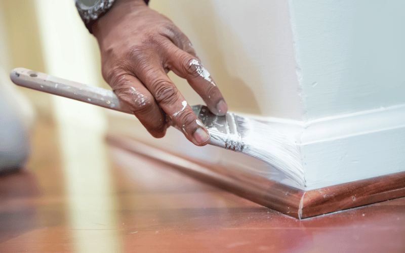 African American male hand holding white paintbrush painting white trim in home over hardwood floor