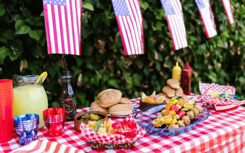 Picnic table with red checkered cloth covered in BBQ food with hanging US flags