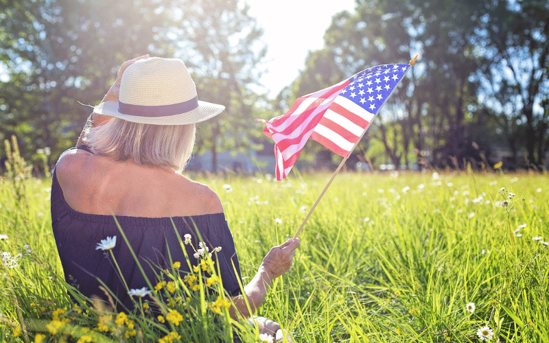 women with short blonde hair wearing white hat with black ribbon and black shirt waving American flag sitting in the grass