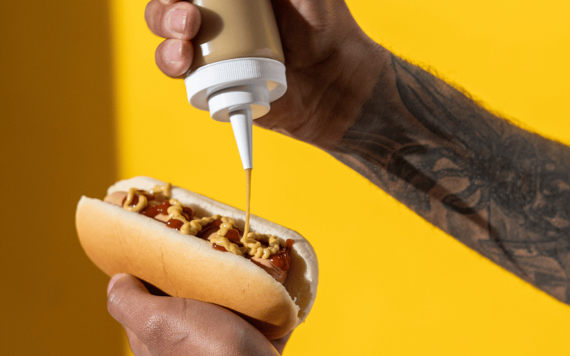 Tattooed male hand holding dog dog squeezing mustard onto it in front of yellow background