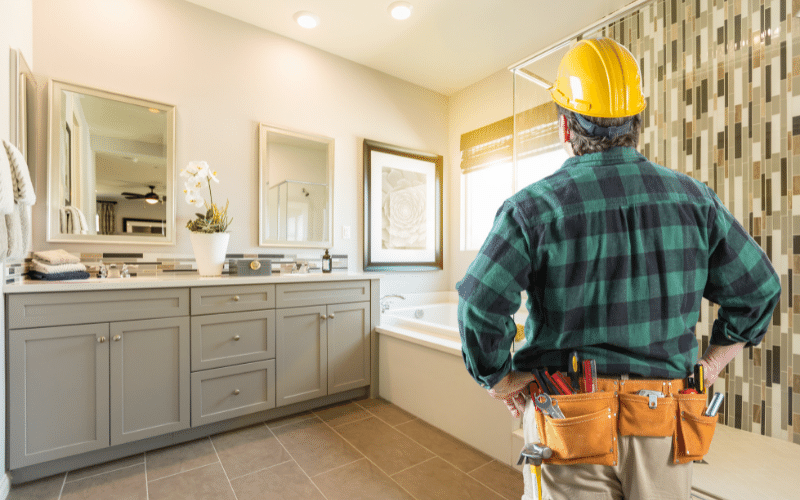 Renovated bathroom with contractor wearing yellow hard hat and blue-green plaid shirt wearing tool belt looking at his good work