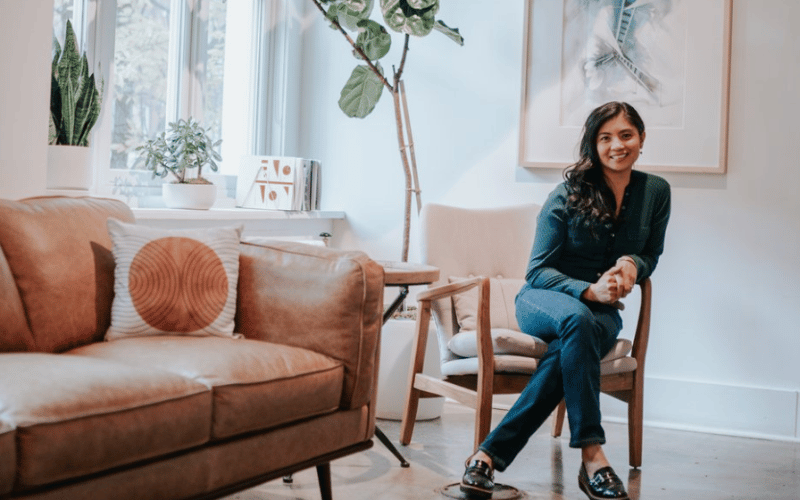 Grace Salaysay wearing dark green blouse and blue jeans sitting in chair beside brown leather couch with houseplants in background smiling