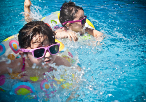 Two little girls with brown hair and pink sunglasses splashing in pool with water rings around them