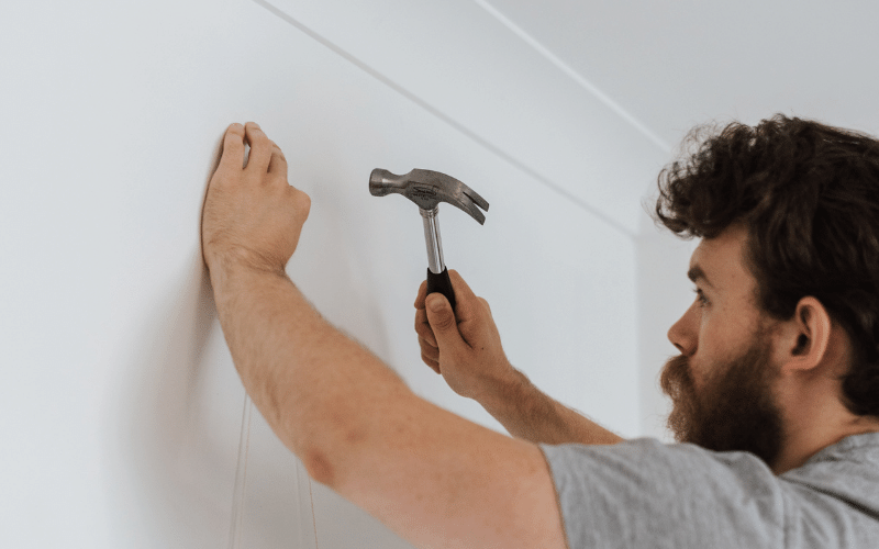 Man with dark hair wearing grey shirt hammering in nail into white wall with right hand