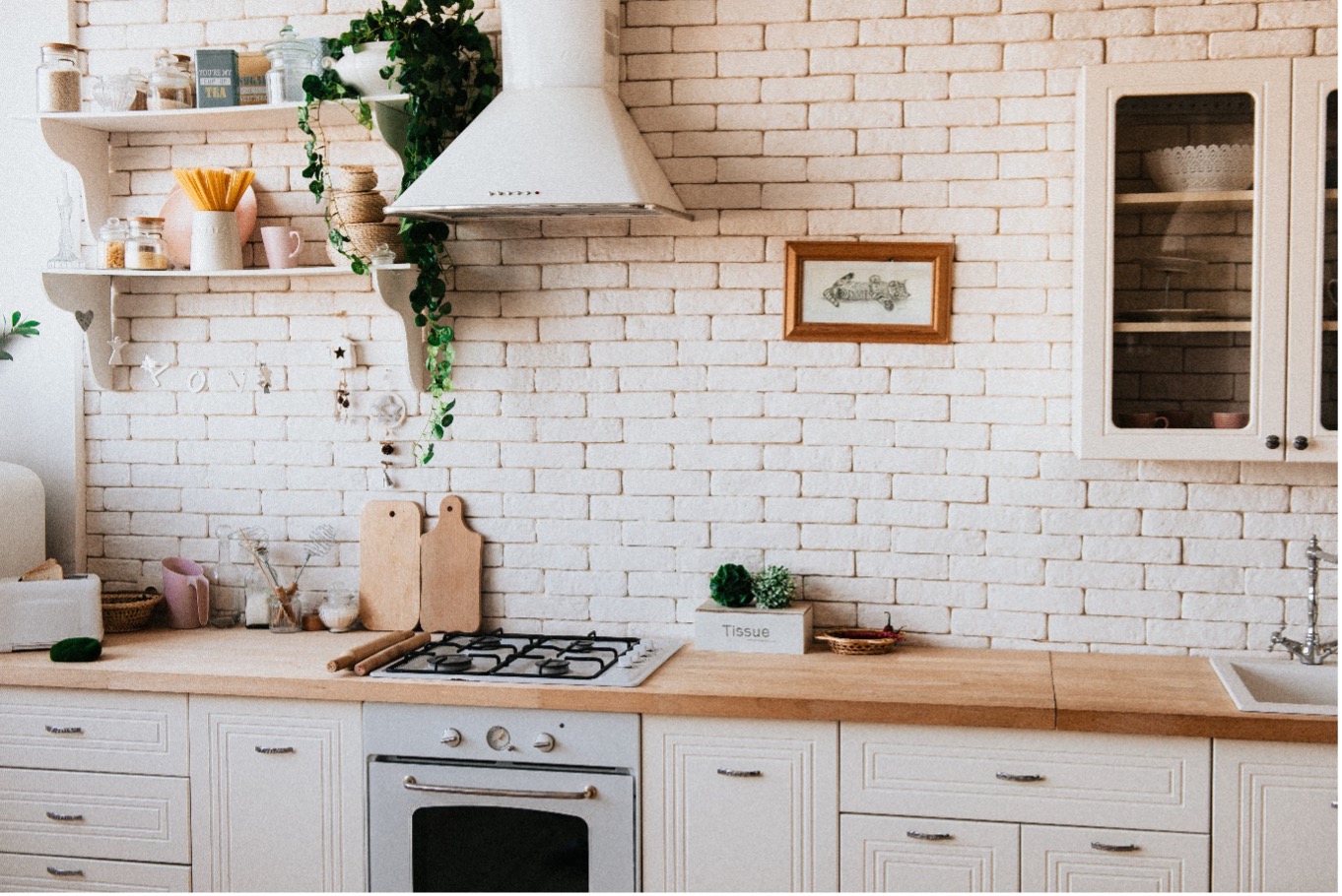 White brick kitchen wall with basic clean wooden countertops with hanging art, shelving and houseplants personalizing the kitchen