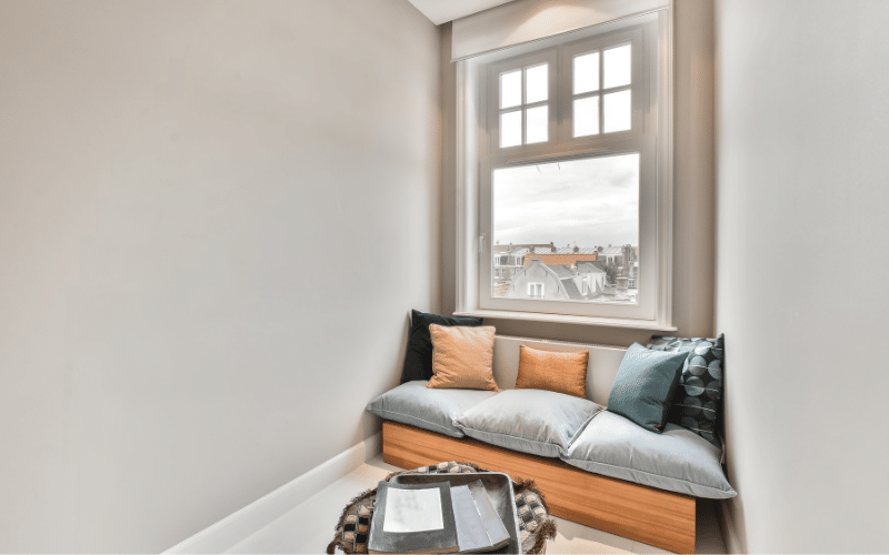 Reading nook in window on second floor with light grey seats, blue and orange pillows and a small table with a TV tray