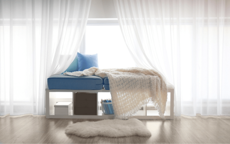 White metal bench in window with bluish purple pillows and white blankets. Storage of boxes and books under the bench with white throw rug in front
