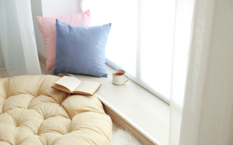 Light pink and purple pillow and cup of tea on white window sill with circular beige plush pillow and open book