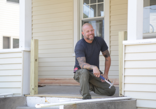 HGTV Joe Mazza kneeling on renovated porch with hammer in left hand