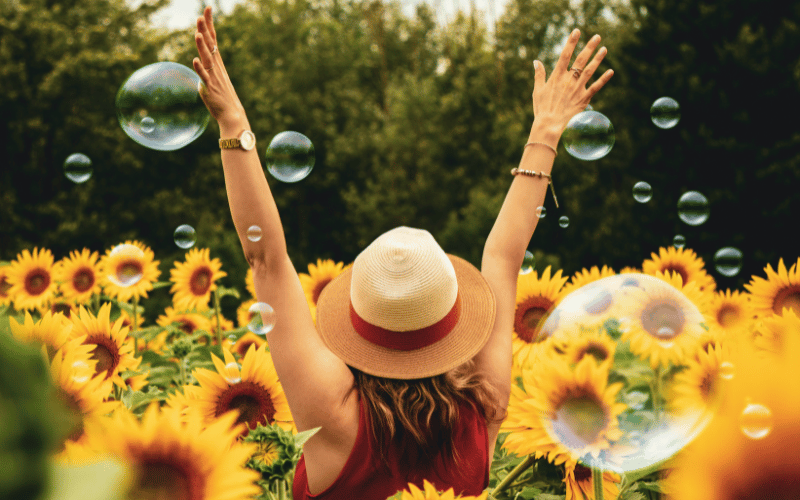 Woman wearing sunhat from back with raised arms walking through sunflower field with bubbles