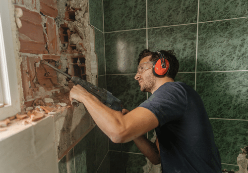 young attractive brown haired white man using jackhammer on bathroom wall wearing orange ear protection